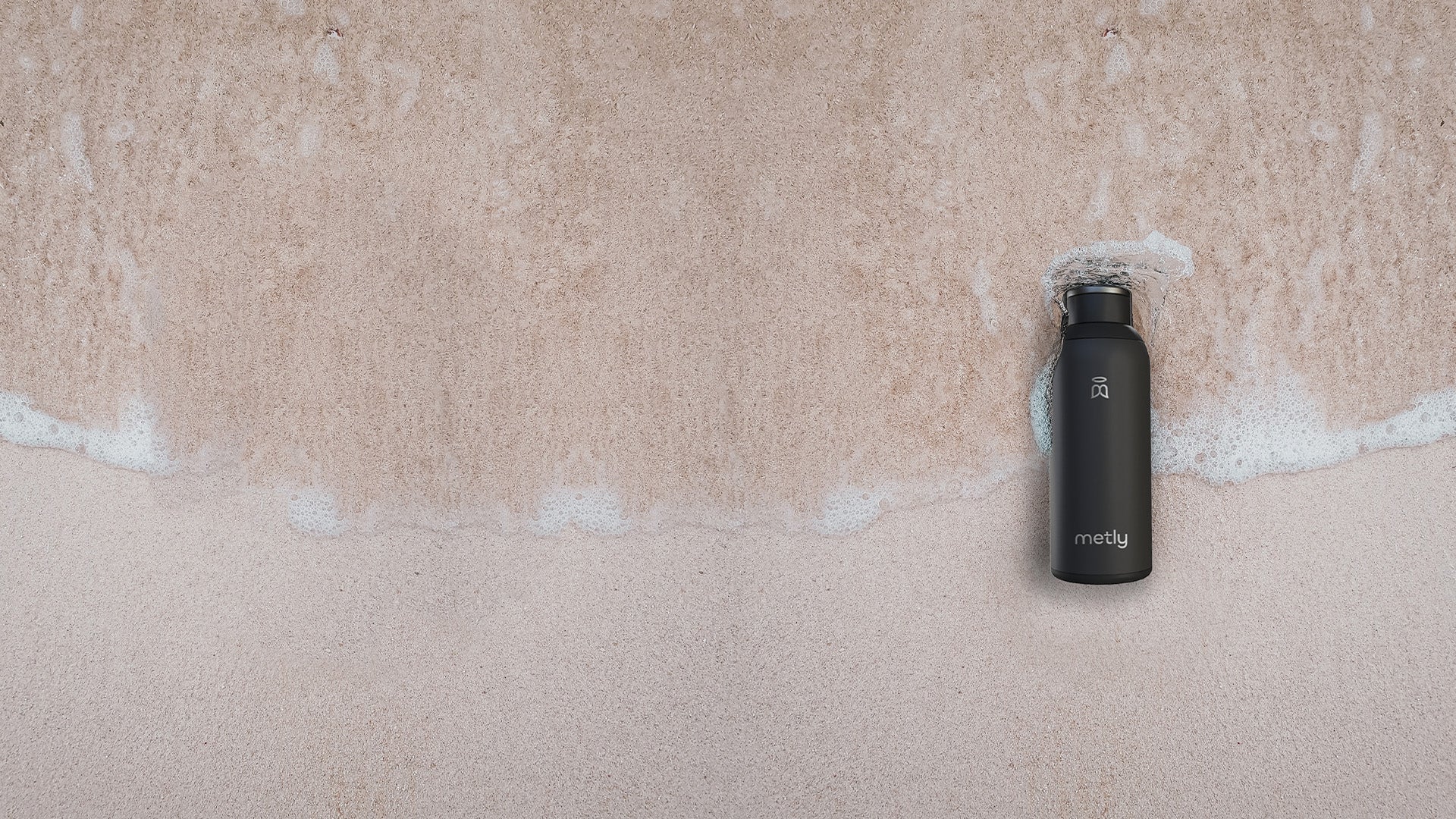 A black metly water bottle set down on a calm beach with the sea water passing over the bottle