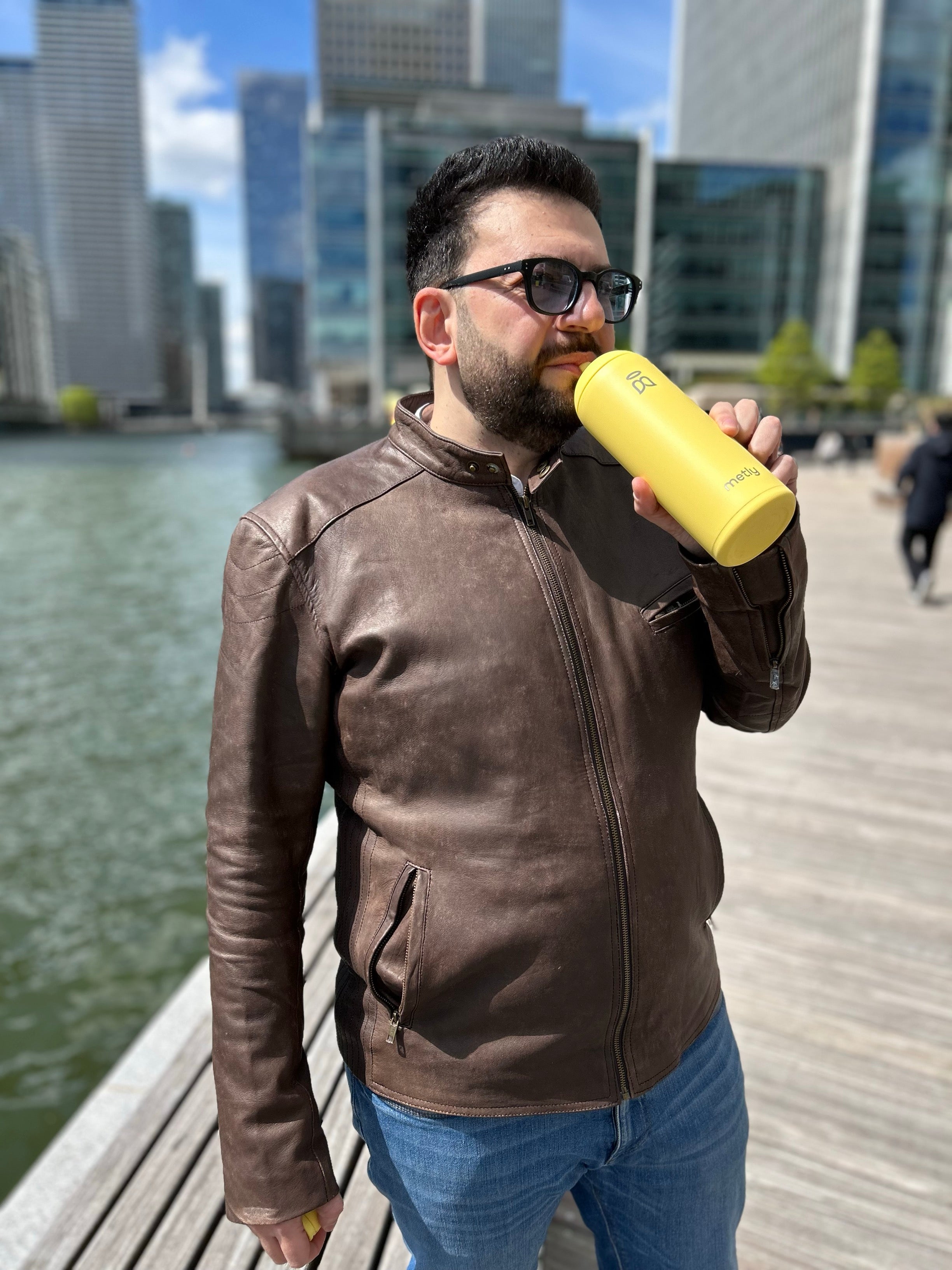 Man in brown leather jacket drinking from a custard yellow 500ml filtered water bottle by a waterfront in the city